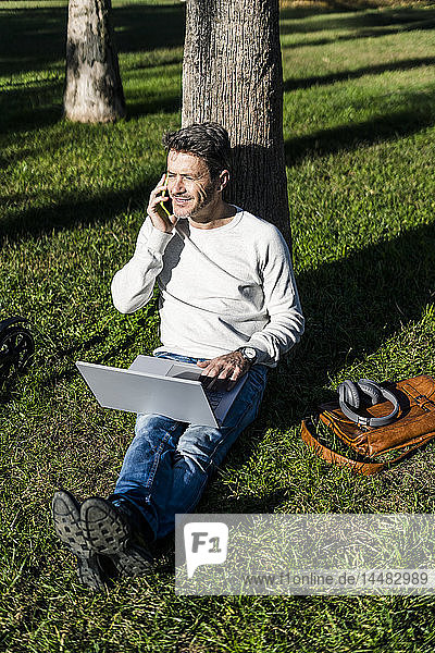 casual businessman sitting on grass in a park  using laptop  talking on the phone