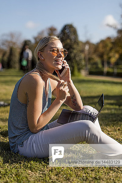 Junge Frau sitzt im Park und benutzt Handy und Laptop