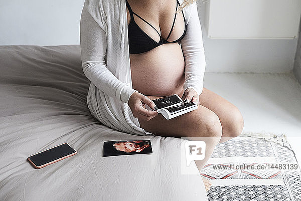 Schwangere Frau zu Hause am Bett sitzend mit Ultraschallbildern