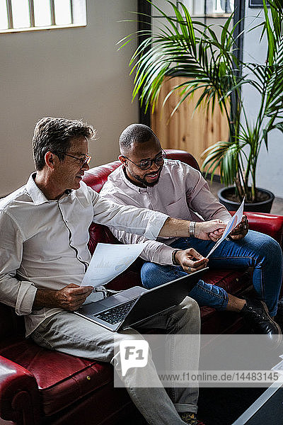 Two businessmen using laptop and discussing documents on sofa in loft office