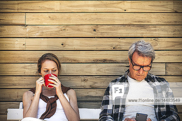 Frau trinkt Kaffee und Mann benutzt Smartphone vor einer Blockhütte