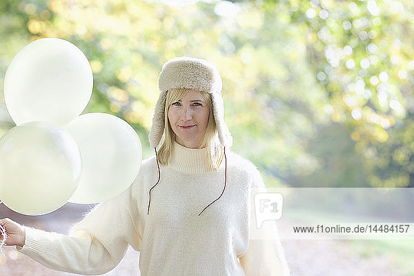 Portrait confident woman in white fur hat holding bunch of balloons in park