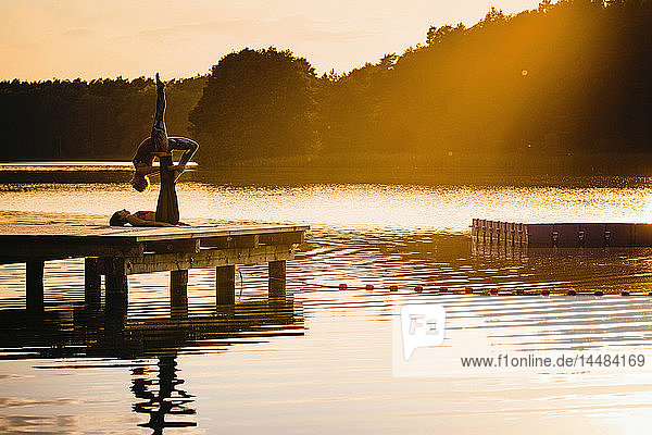 Frauen üben Acro-Yoga auf einem Dock am See bei Sonnenuntergang