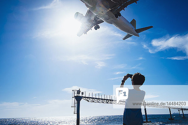 Boy with camera phone photographing airplane flying low overhead near Lanzarote Airport