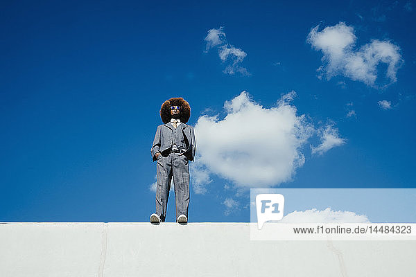 Selbstbewusster  cooler  gut gekleideter junger Mann mit Afro  der an einer sonnigen Wand vor blauem Himmel steht