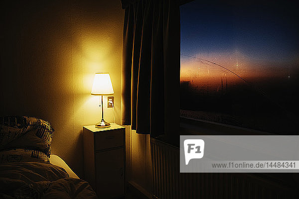 Nachttischlampe  die die Schlafzimmerecke neben dem Fenster mit Blick auf den Abendhimmel beleuchtet