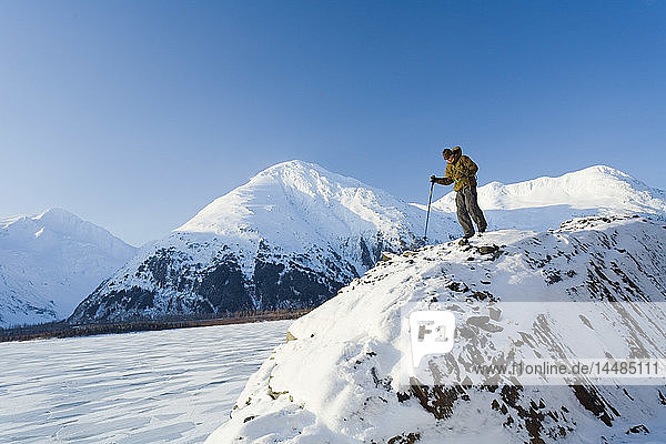 Ein Bergsteiger steht in der Nähe der Spitze einer Klippe über der gefrorenen Oberfläche des Portage Lake im Chugach National Forest. Winter in Süd-Zentral-Alaska.
