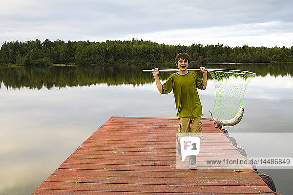 Junger Junge hält einen Coho-Lachs in einem Fischernetz auf einem Steg an einem See in Anchorage im Sommer in Süd-Zentral-Alaska