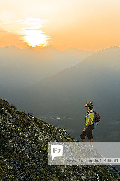 Hiker on the steep hillside of Mt. Alyeska watching the sun set over Girdwood Valley in Southcentral Alaska