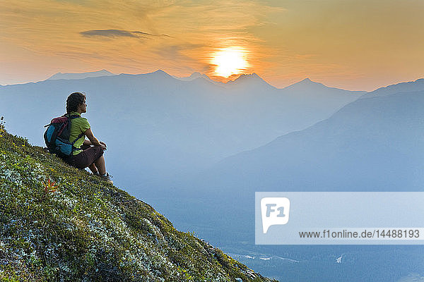 Hiker sits on steep hillside of Mt. Alyeska watching the sun set over Girdwood Valley in Southcentral Alaska