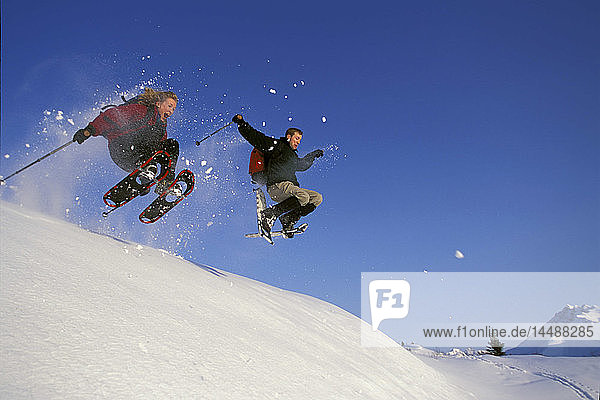 Ehepaar springt mit Schneeschuhen von Felsvorsprung Arctic Valley AK Winter Southcentral