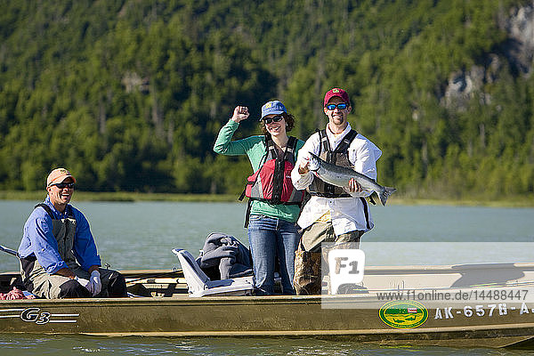 Mann und Frau stehen im Boot und halten den Lachsfang des Tages auf dem Big River Lakes mit einem Angelführer und den Chigmit Mountains im Hintergrund während des Sommers in Southcentral Alaska