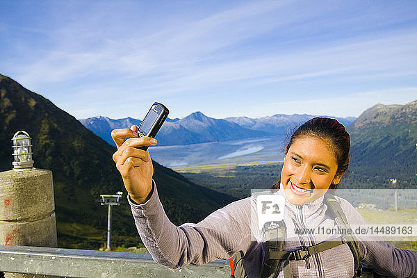 Young adult Hispanic woman takes a picture of herself using a cell phone on top of Mt. Alyeska with view of Chugach Mountains near Girdwood in Southcentral Alaska