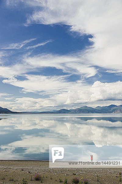 Eine Frau steht am Ufer des Kluane Lake mit Bergen und Wolken  die sich im ruhigen Wasser des Kluane Lake spiegeln  Yukon Territorium  Kanada  Sommer