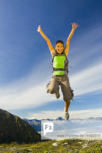 Junge erwachsene hispanische Frau springt vor Freude auf dem Gipfel des Berges Alyeska mit Blick auf die Chugach Mountains in der Nähe von Girdwood in Südalaska