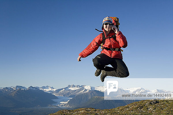 Männlicher Wanderer mit Handy  der auf einem Bergrücken oberhalb der Inside Passage im Sommer in Alaska aufgeregt herumspringt