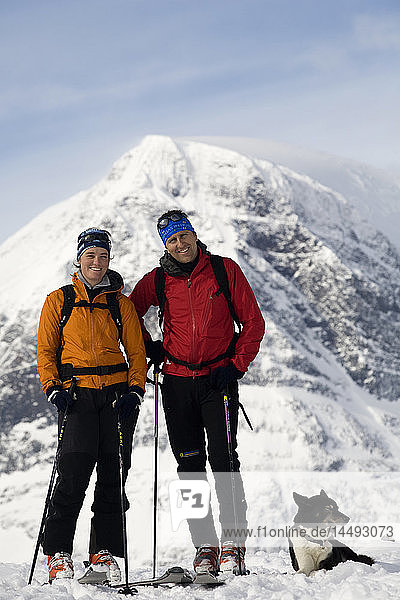 Two people standing with dog near snowcapped mountain
