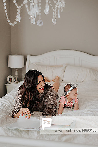 Mother reading with baby daughter on bed in bedroom