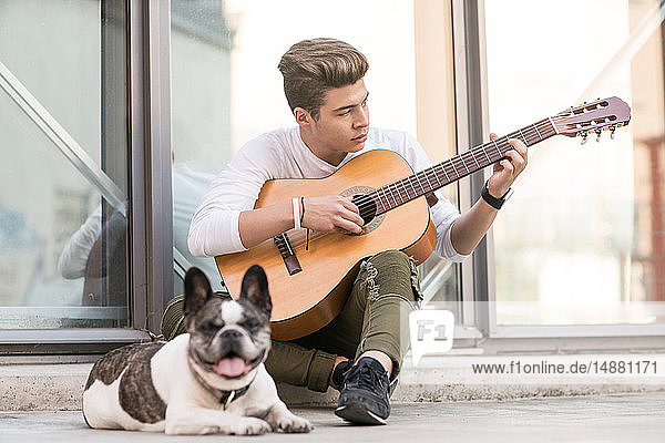 Jugendlicher Flüchtlingsjunge spielt mit Hund Gitarre auf dem Bürgersteig