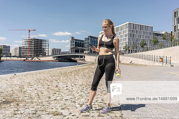 Young woman taking break from exercise and using smartphone in city  Berlin  Germany