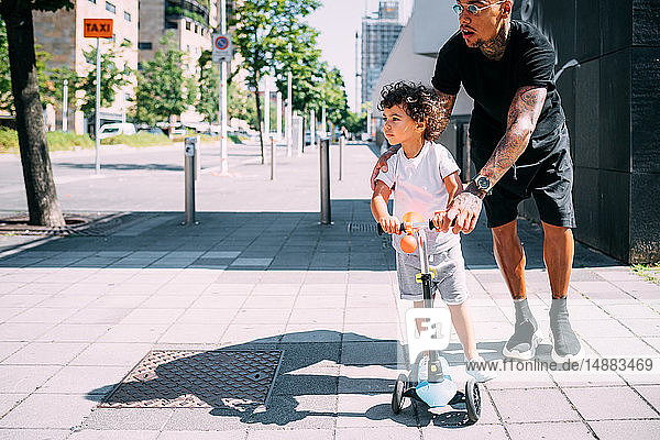Father teaching son ride push scooter on sidewalk