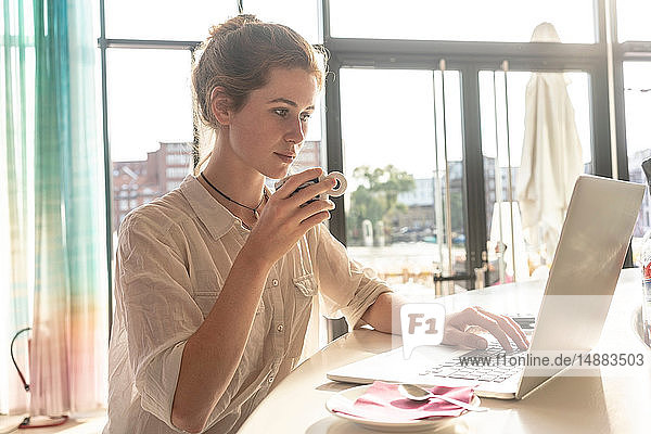 Junge Frau benutzt Laptop und trinkt an sonnigen Tagen Kaffee im Büro