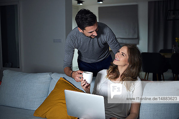 Junger Mann gibt seiner abends auf dem Sofa sitzenden Freundin Kaffee