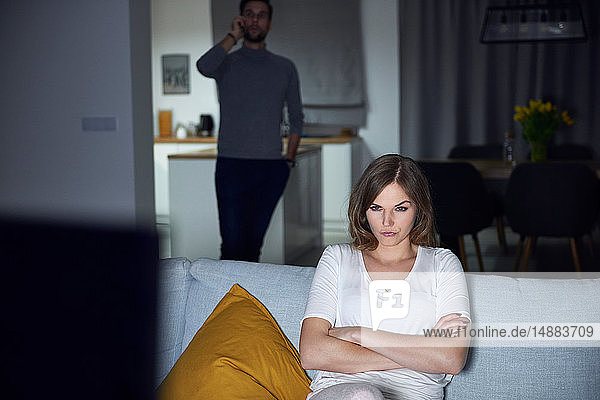Junge Frau schmollt abends auf dem Sofa  während ihr Freund mit dem Smartphone telefoniert