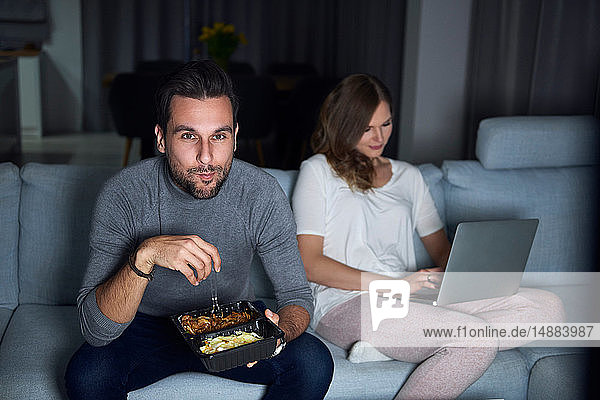 Junges Paar sitzt abends auf dem Sofa  isst zum Mitnehmen und benutzt Laptop