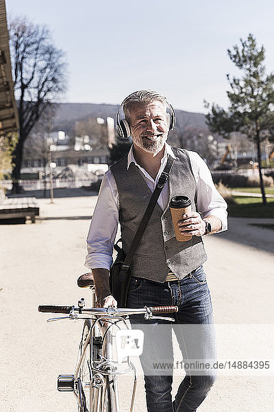 Lächelnder reifer Geschäftsmann mit Fahrrad  Kaffee zum Mitnehmen und Kopfhörern unterwegs in der Stadt
