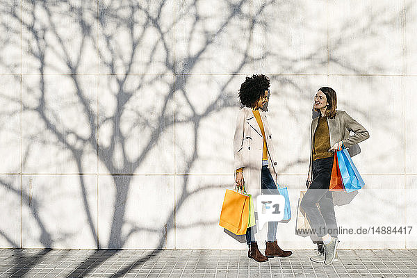 Two happy women with shopping bags standing at a wall talking