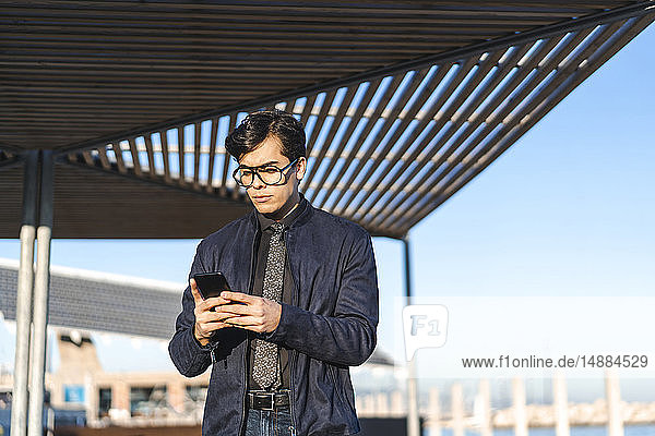 Porträt eines modischen jungen Geschäftsmannes mit Brille und Krawatte beim Blick auf das Mobiltelefon