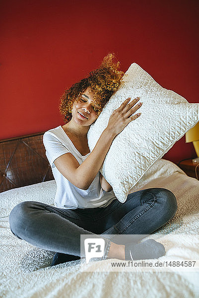 Junge Frau mit lockigem Haar sitzt zu Hause auf dem Bett und hält Kissen