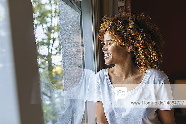 Lächelnde junge Frau mit lockigem Haar  die zu Hause aus dem Fenster schaut