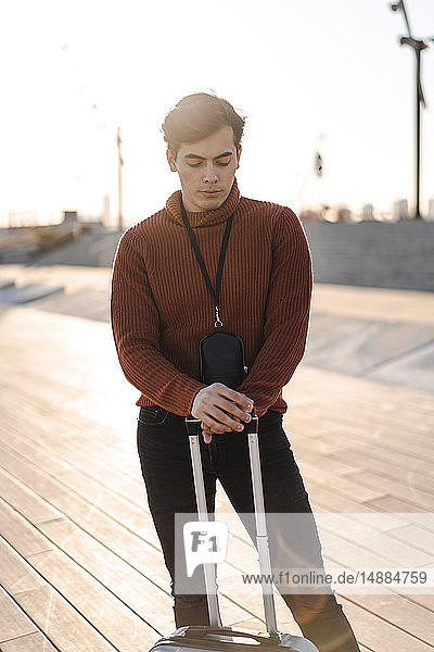Portrait of young fashionable man with luggage standing at backlight