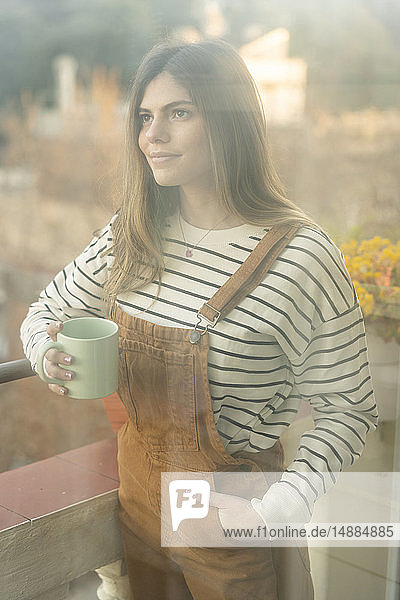 Porträt einer lächelnden jungen Frau mit Kaffeetasse auf dem Balkon stehend
