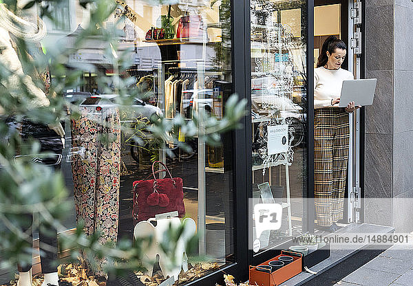 Shop owner standing in door of fashion store  using laptop