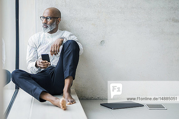 Barefoot mature businessman with smartphone sitting on window sill looking at distance