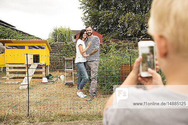 Junge macht Handyfoto von Eltern  die im Hühnerstall im Garten stehen