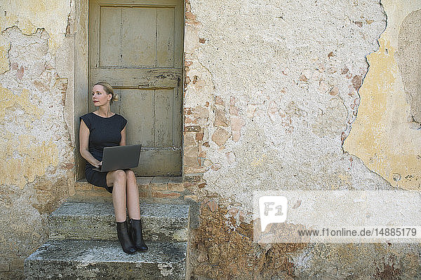 Italien  Toskana  Monteriggioni  Frau sitzt mit Laptop am Hauseingang