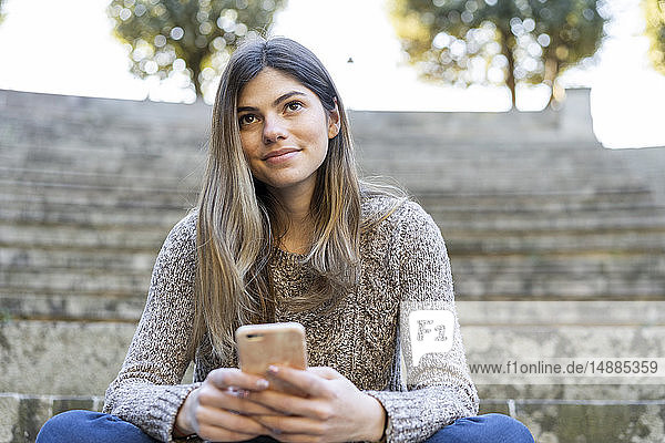 Junge Frau sitzt auf einer Treppe im Freien und hält ein Mobiltelefon