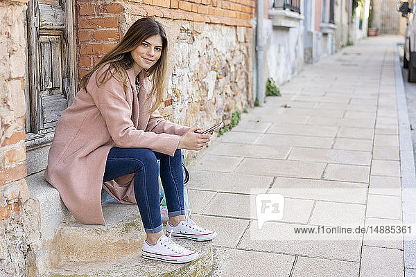 Porträt einer jungen Frau  die am Hauseingang sitzt und ein Handy in der Hand hält