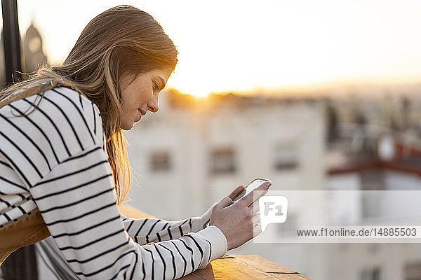 Junge Frau auf Balkon bei Sonnenuntergang mit Mobiltelefon