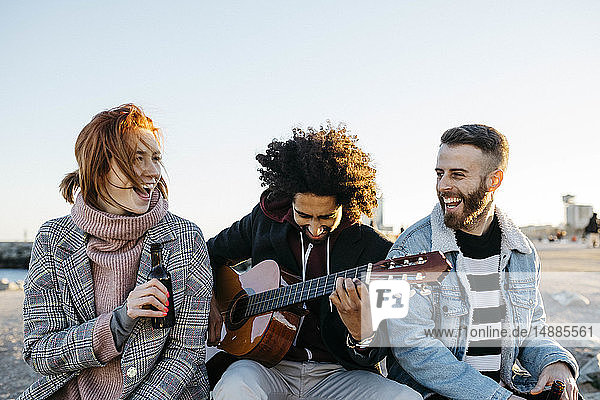 Drei glückliche Freunde mit Gitarre sitzen bei Sonnenuntergang im Freien