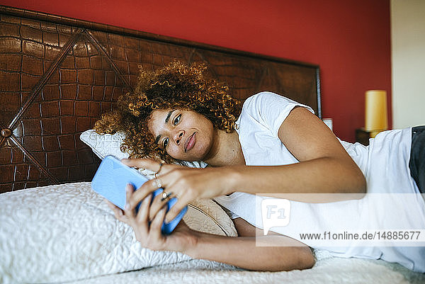 Junge Frau mit lockigem Haar liegt zu Hause im Bett und benutzt ihr Handy