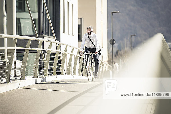 Lächelnder reifer Geschäftsmann beim Fahrradfahren auf einer Brücke in der Stadt