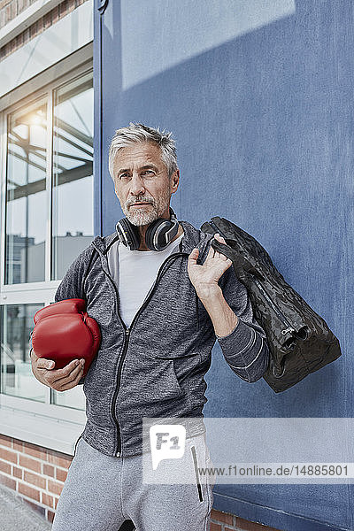 Porträt eines reifen Mannes mit Kopfhörer  Sporttasche und roten Boxhandschuhen vor der Turnhalle stehend