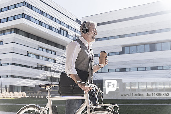 Smiling mature businessman with bicycle  takeaway coffee and headphones on the go in the city