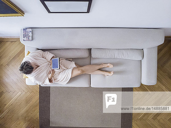 Reife Frau im Bademantel sitzt zu Hause auf der Couch und benutzt ein digitales Tablet