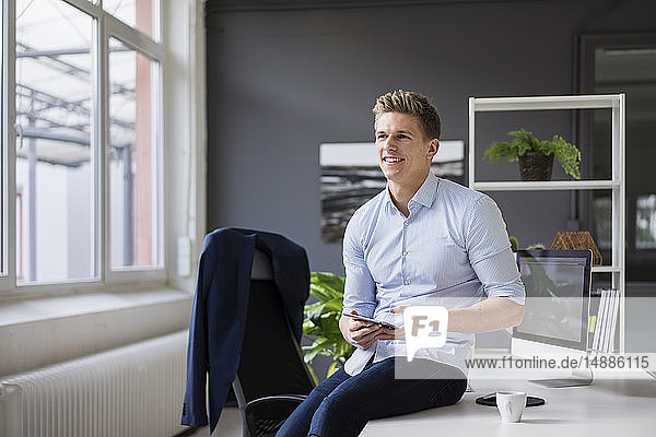 Smiling young businessman sitting on desk in office holding tablet